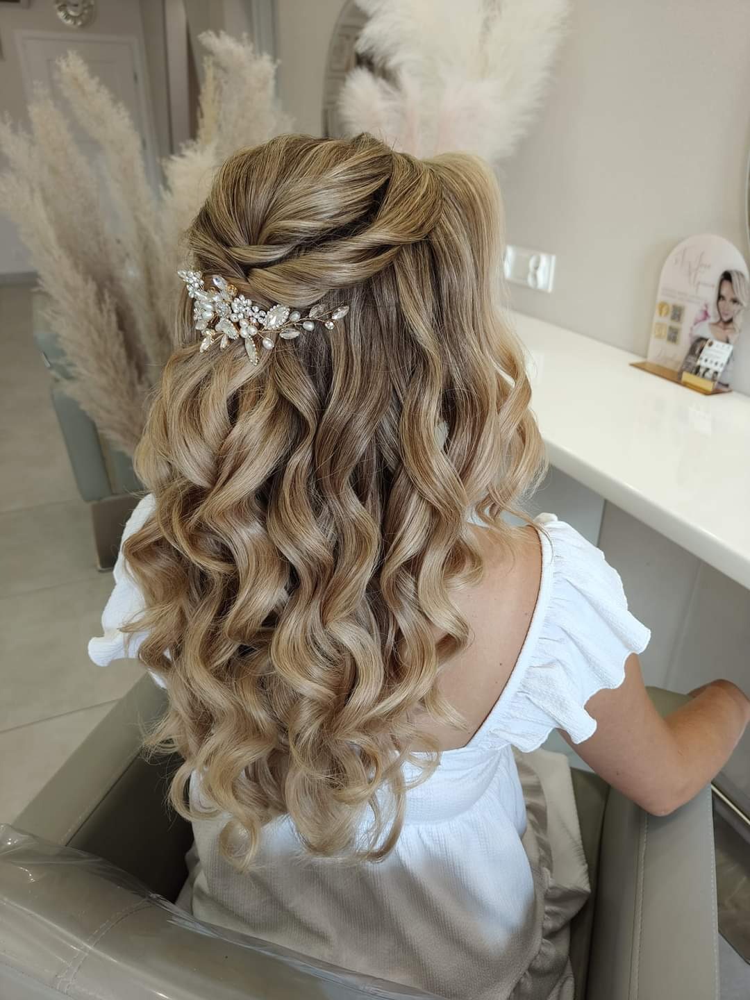 Upięcie ślubne z loków, ozdobione spinką z kryształkami i perłami, widok z tyłu na blond włosy w salonie fryzjerskim.