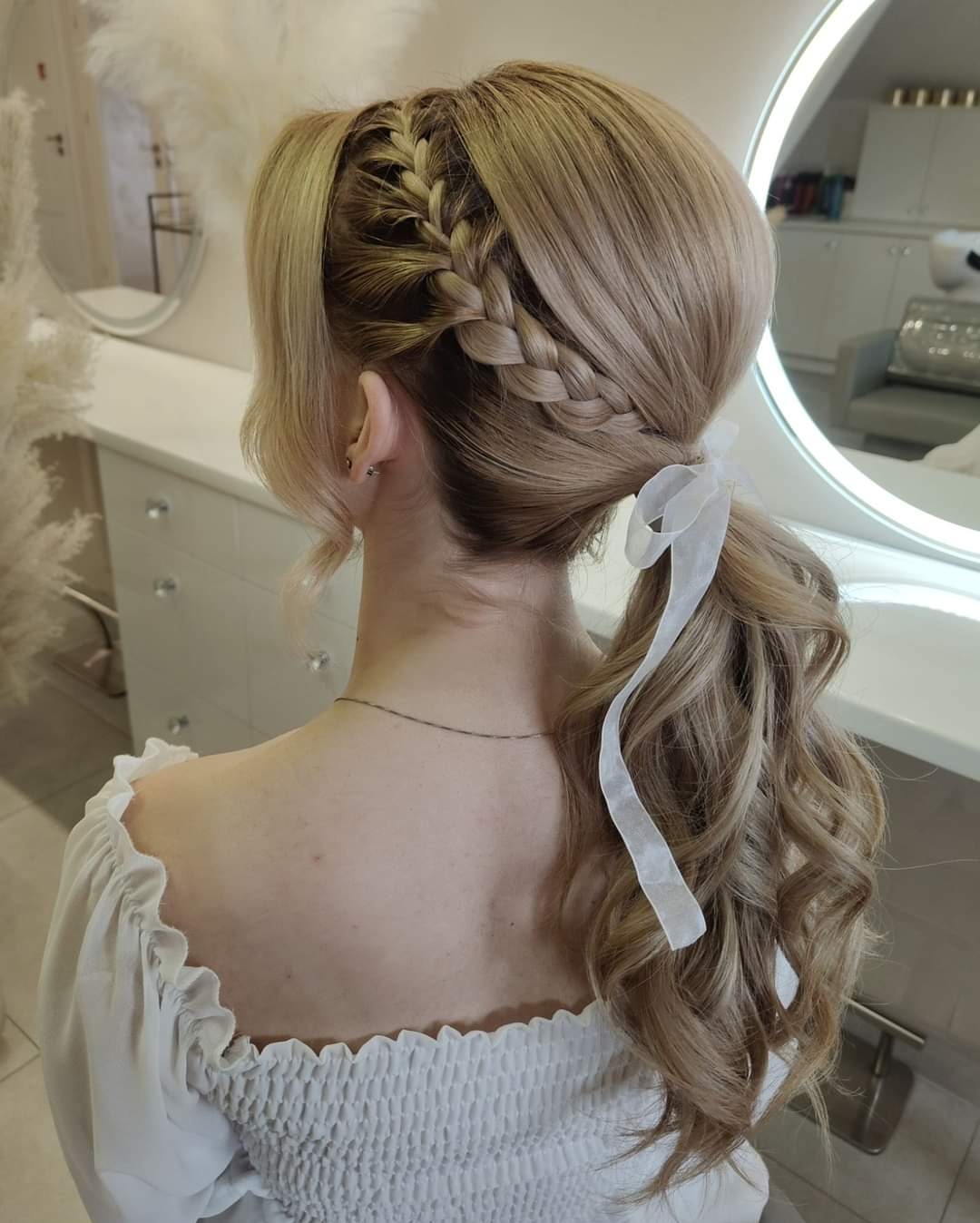 Blond włosy upięte w wysoki kucyk z warkoczem i białą wstążką, widok z tyłu na tle jasnego wnętrza salonu.