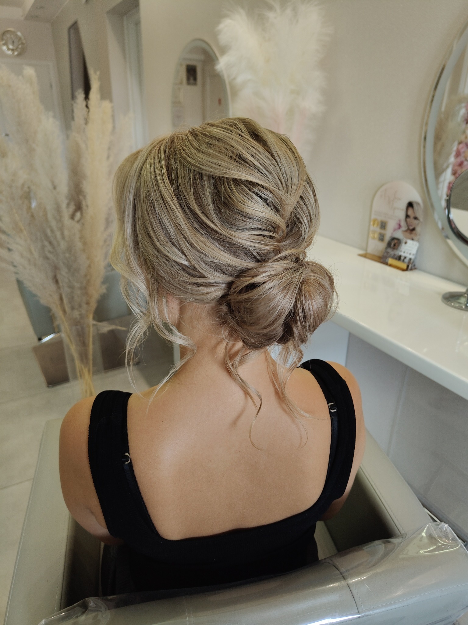 Upięcie włosów blond w kok na karku, widok z tyłu, w tle dekoracyjne trawy i toaletka z lustrem.