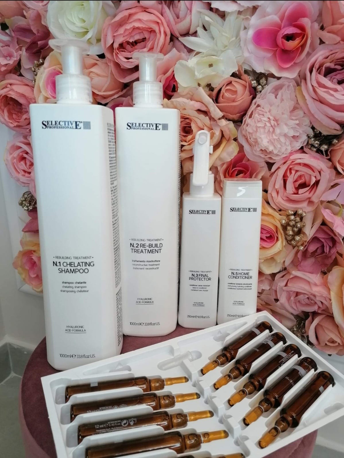Zestaw kosmetyków do włosów Selective Professional: szampon chelatujący, odbudowujący treatment, final protector i home conditioner, oraz ampułki w białym opakowaniu, na tle bukietu różowych róż.