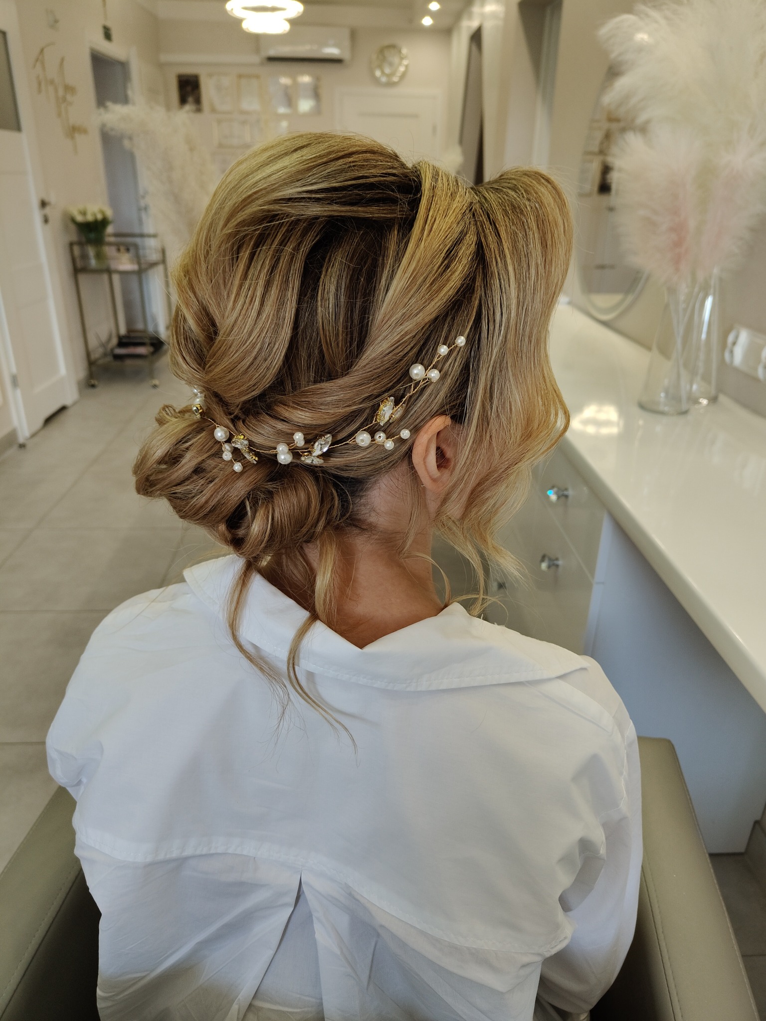 Upięcie ślubne z blond włosów z ozdobną spinką z pereł i kamieni, widok z tyłu, biała koszula, jasne wnętrze salonu.