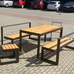 Stal Projekt - Zestaw mebli ogrodowych: stół z blatem z jasnego drewna oraz dwie ławki i krzesło, wszystkie z czarną, metalową ramą, ustawione na brukowanym placu parkingowym.