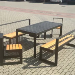 Zestaw mebli miejskich: stół z blatem z ciemnych listew i dwie ławki z oparciami oraz krzesło z drewnianymi siedziskami i czarną stalową ramą, ustawione na kostce brukowej przed budynkiem...