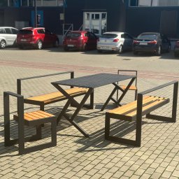 Stal Projekt - Zestaw mebli ogrodowych: stół z blatem z czarnych listew i dwie ławki z oparciami z jasnego drewna, na tle parkingu z samochodami i szarego budynku.