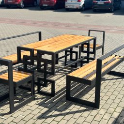 Stal Projekt - Nowoczesny zestaw mebli ogrodowych z drewna i czarnego metalu, składający się ze stołu i dwóch ławek z oparciami, ustawiony na brukowanym placu, z zaparkowanymi samochodami w tle.