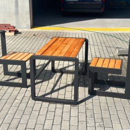 Stal Projekt - Zestaw mebli ogrodowych: stół z blatem z desek i dwa krzesła z siedziskami z desek, czarna metalowa rama o kwadratowym przekroju, na kostce brukowej, w tle brama garażowa.