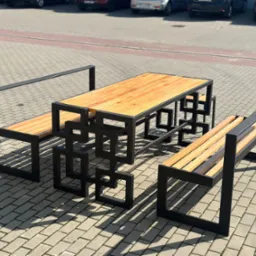 Zestaw mebli ogrodowych: stół z blatem z jasnego drewna i dwie ławki z oparciami, charakteryzujące się nowoczesną, geometryczną konstrukcją z czarnych, stalowych profili. Całość ustawiona na szarej...