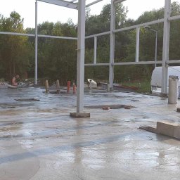 Stal Projekt - Wylana, mokra betonowa posadzka hali w trakcie budowy, widoczne stalowe słupy konstrukcyjne, elementy instalacji i pracujący w tle mężczyzna.