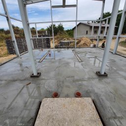 Stal Projekt - Wnętrze budowanej hali z widoczną konstrukcją stalową, mokrą betonową posadzką i deską na pierwszym planie, w tle widoczny budynek i teren.