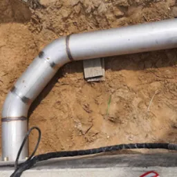 Spawany, nierdzewny element rurowy z kolankiem, wkopany w ziemię, Zawiercie. Widoczne spawy i oznaczenia producenta na rurze.