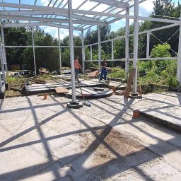 Stal Projekt - Wnętrze budowanej hali z widoczną stalową konstrukcją szkieletową, betonową posadzką w trakcie prac i dwoma pracownikami w tle.