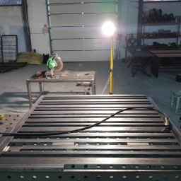 Stal Projekt - Konstrukcja metalowa z profili, prawdopodobnie rama, na stole spawalniczym w warsztacie, oświetlona lampą roboczą, z widocznymi narzędziami do obróbki metalu.