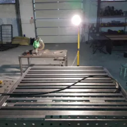Konstrukcja metalowa z profili, prawdopodobnie rama, na stole spawalniczym w warsztacie, oświetlona lampą roboczą, z widocznymi narzędziami do obróbki metalu.