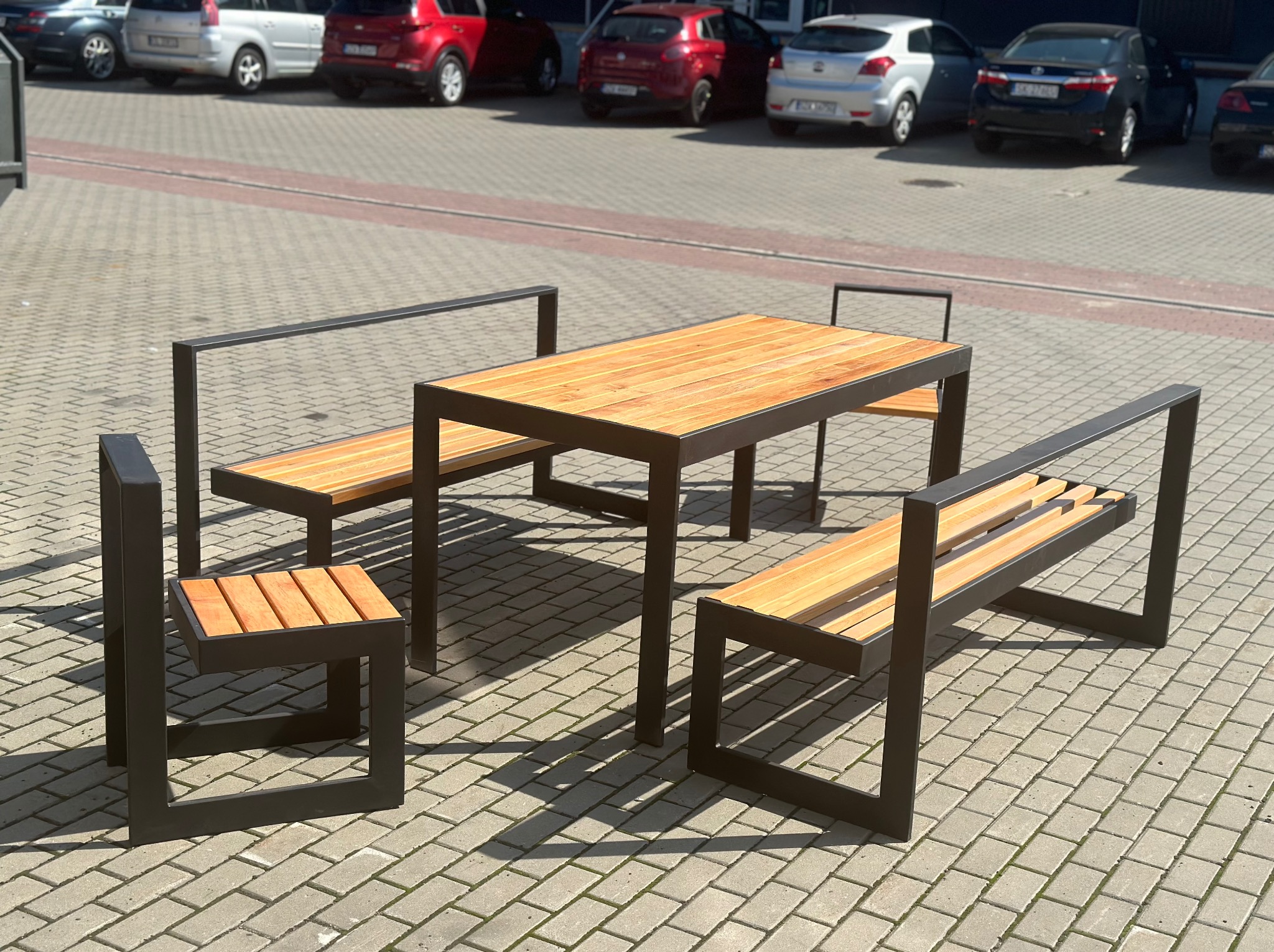 Zestaw mebli ogrodowych: stół z blatem z jasnego drewna oraz dwie ławki i krzesło, wszystkie z czarną, metalową ramą, ustawione na brukowanym placu parkingowym.