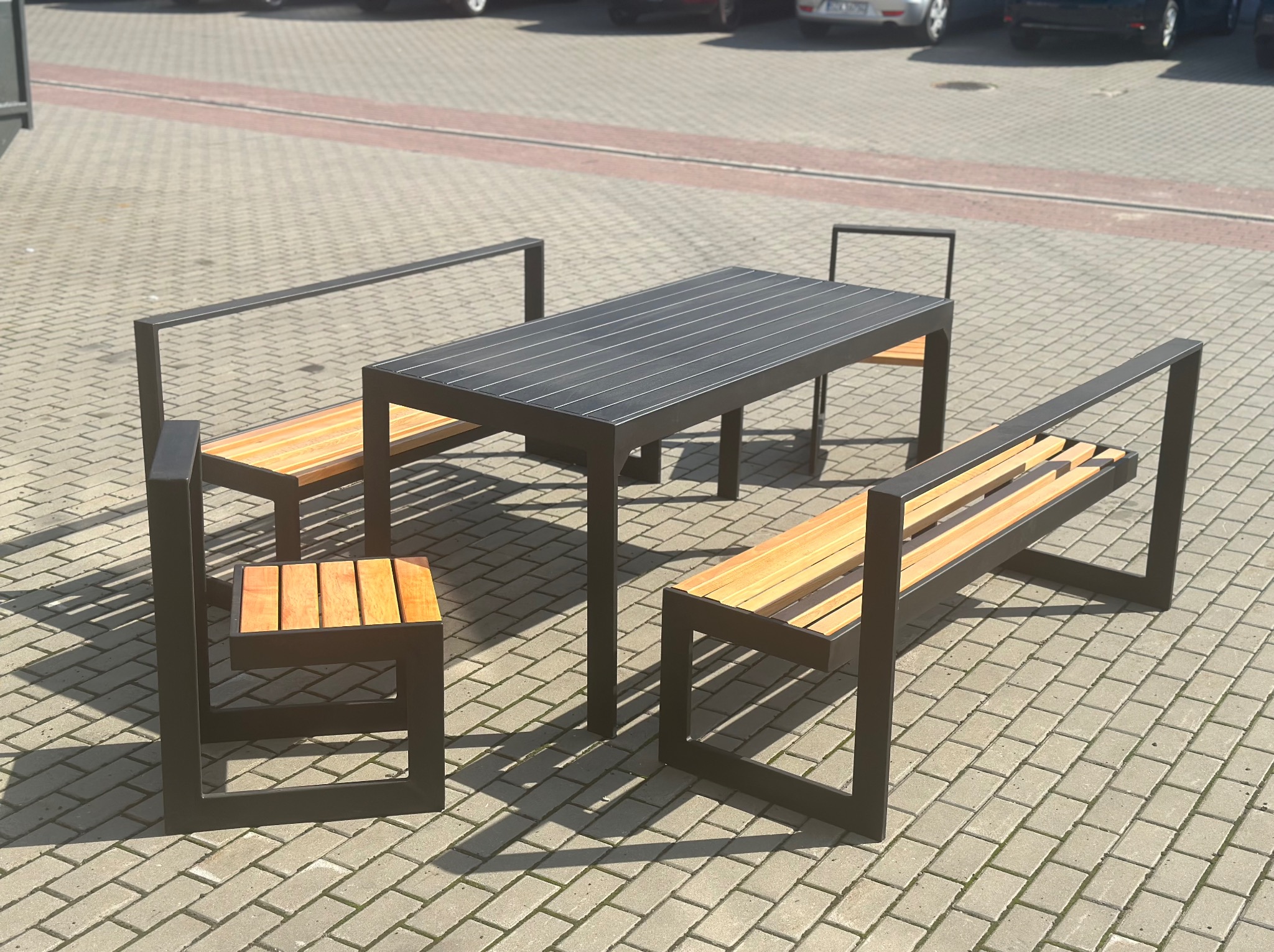 Zestaw mebli miejskich: stół z blatem z ciemnych listew i dwie ławki z oparciami oraz krzesło z drewnianymi siedziskami i czarną stalową ramą, ustawione na kostce brukowej przed budynkiem...