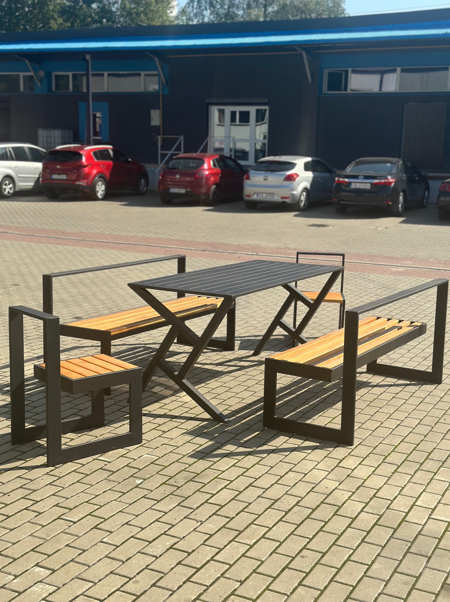 Zestaw mebli ogrodowych: stół z blatem z czarnych listew i dwie ławki z oparciami z jasnego drewna, na tle parkingu z samochodami i szarego budynku.