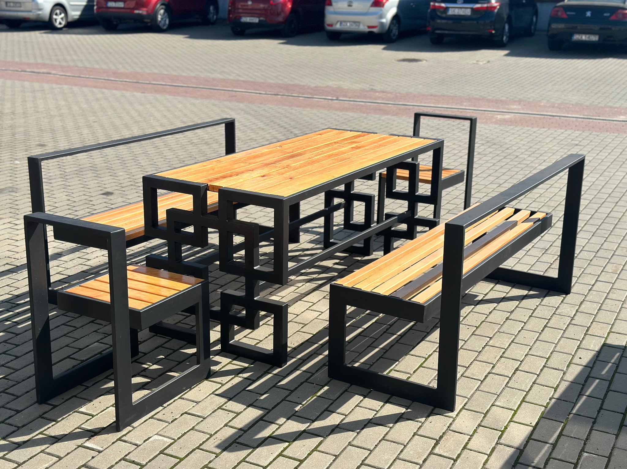 Nowoczesny zestaw mebli ogrodowych z drewna i czarnego metalu, składający się ze stołu i dwóch ławek z oparciami, ustawiony na brukowanym placu, z zaparkowanymi samochodami w tle.
