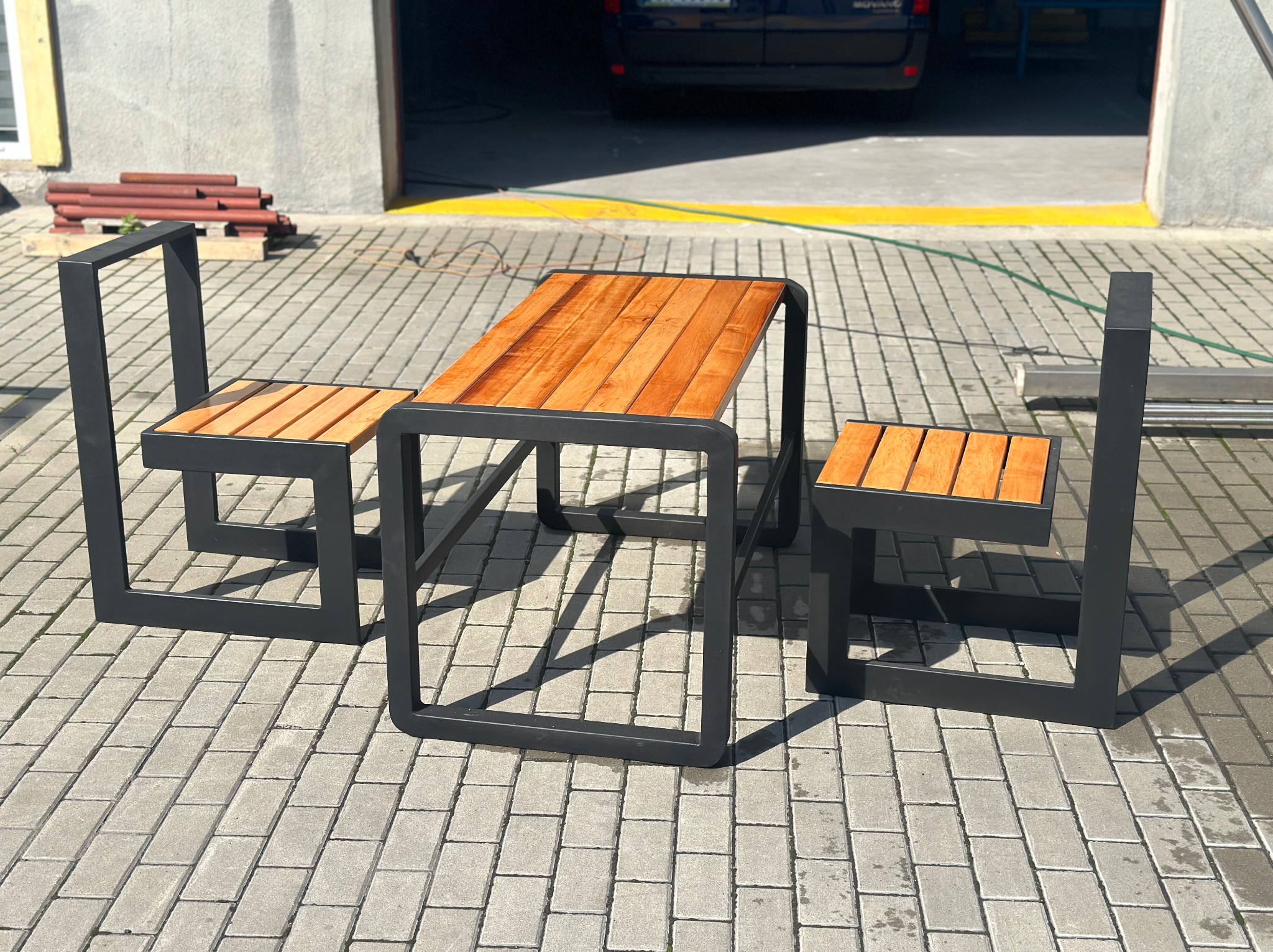 Zestaw mebli ogrodowych: stół z blatem z desek i dwa krzesła z siedziskami z desek, czarna metalowa rama o kwadratowym przekroju, na kostce brukowej, w tle brama garażowa.