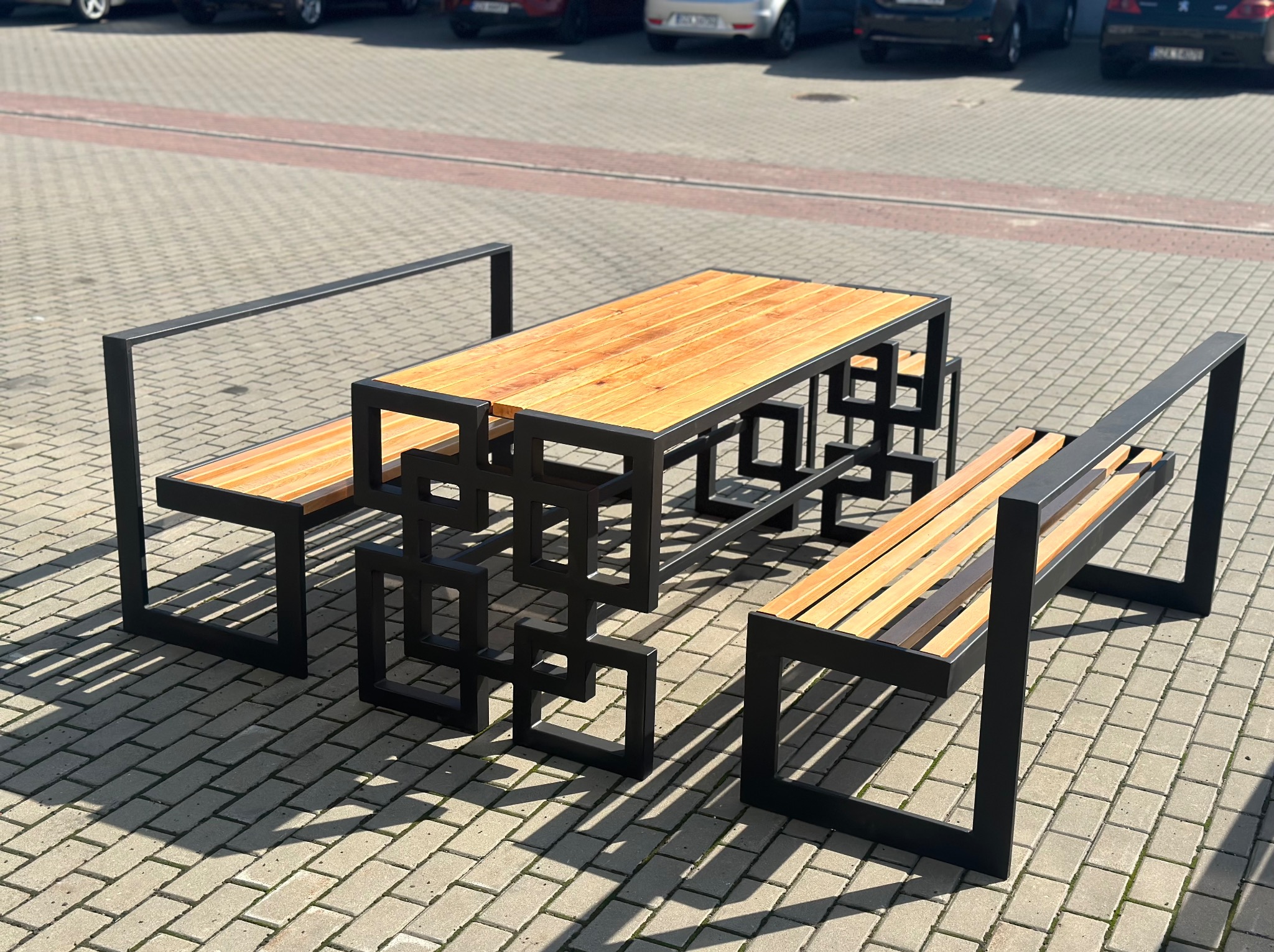 Zestaw mebli ogrodowych: stół z blatem z jasnego drewna i dwie ławki z oparciami, charakteryzujące się nowoczesną, geometryczną konstrukcją z czarnych, stalowych profili. Całość ustawiona na szarej...