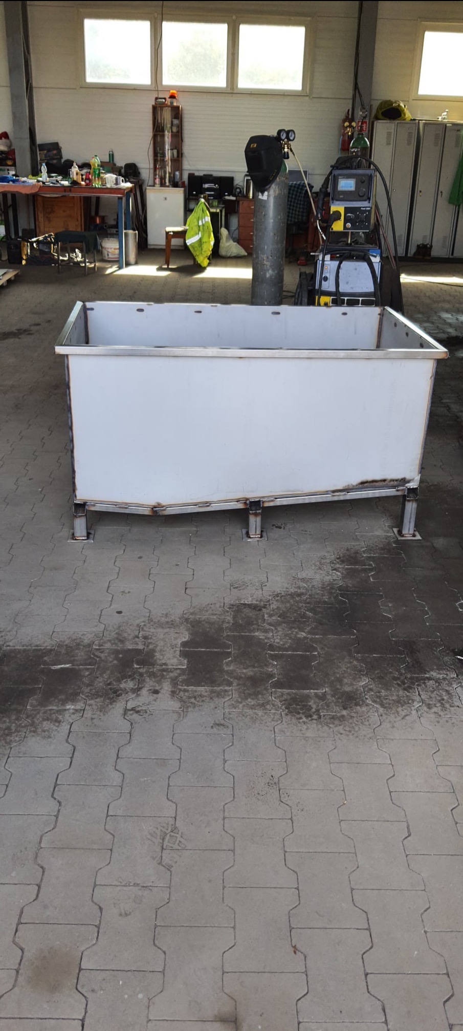 Biały, metalowy zbiornik na czterech nogach, spawany, widoczny w warsztacie z butlą spawalniczą i sprzętem w tle.