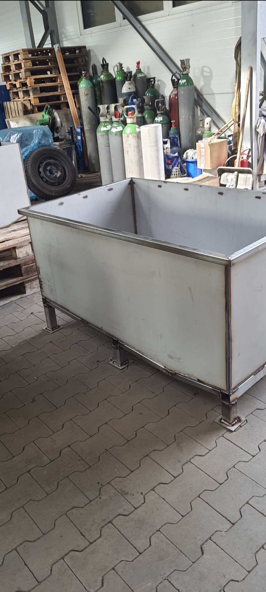 Metalowy kontener spawalniczy na czterech nogach, widoczne spawy, w tle butle z gazem i palety.