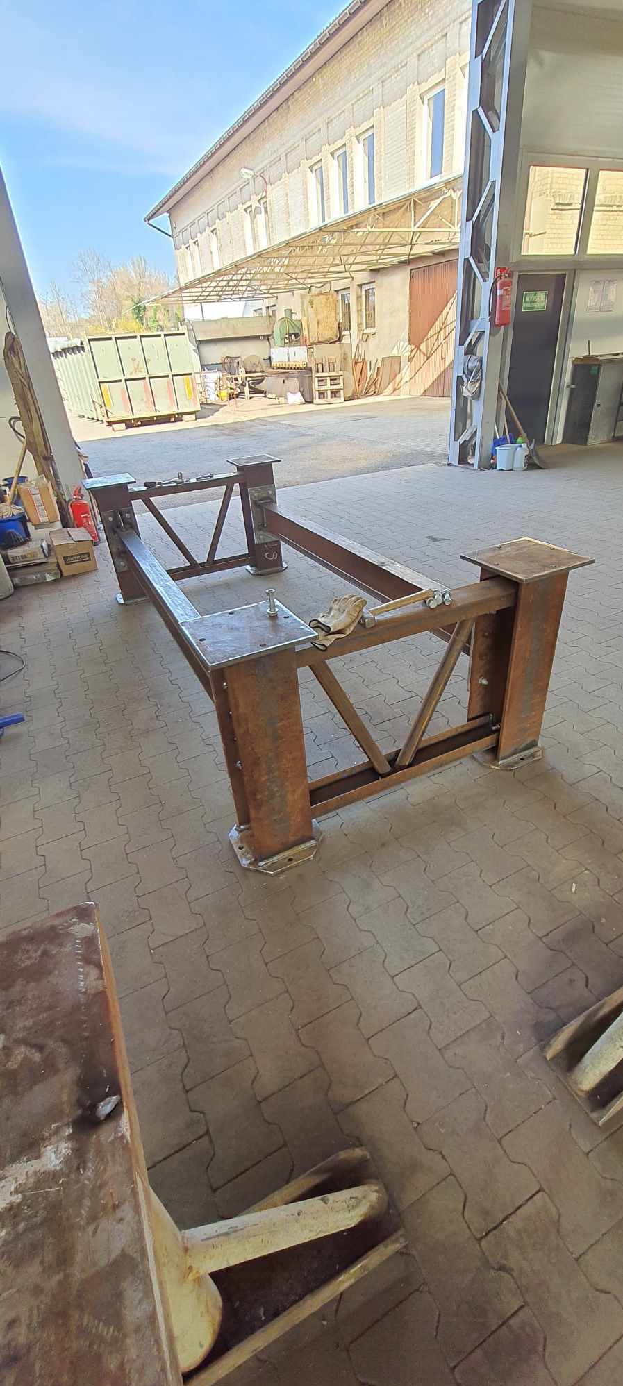 Konstrukcja stalowa w trakcie budowy w hali, widoczne elementy spawane, surowe wykończenie, na podłodze leżą rękawice robocze.