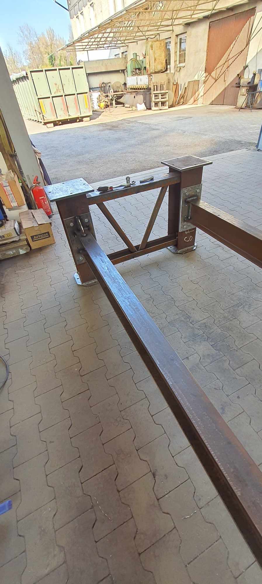 Konstrukcja stalowa z profili, spawana, częściowo zmontowana, widoczne narzędzia i elementy mocujące, tło warsztatowe z kontenerem i sprzętem.