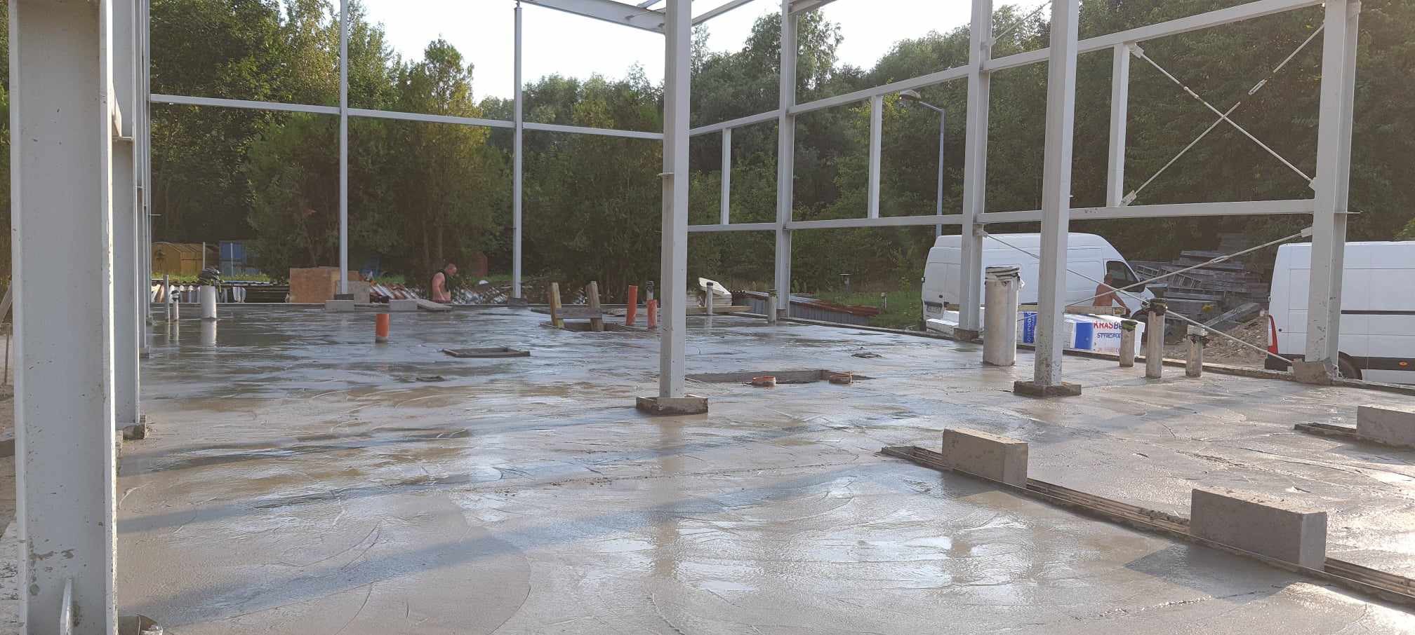 Wylana, mokra betonowa posadzka hali w trakcie budowy, widoczne stalowe słupy konstrukcyjne, elementy instalacji i pracujący w tle mężczyzna.