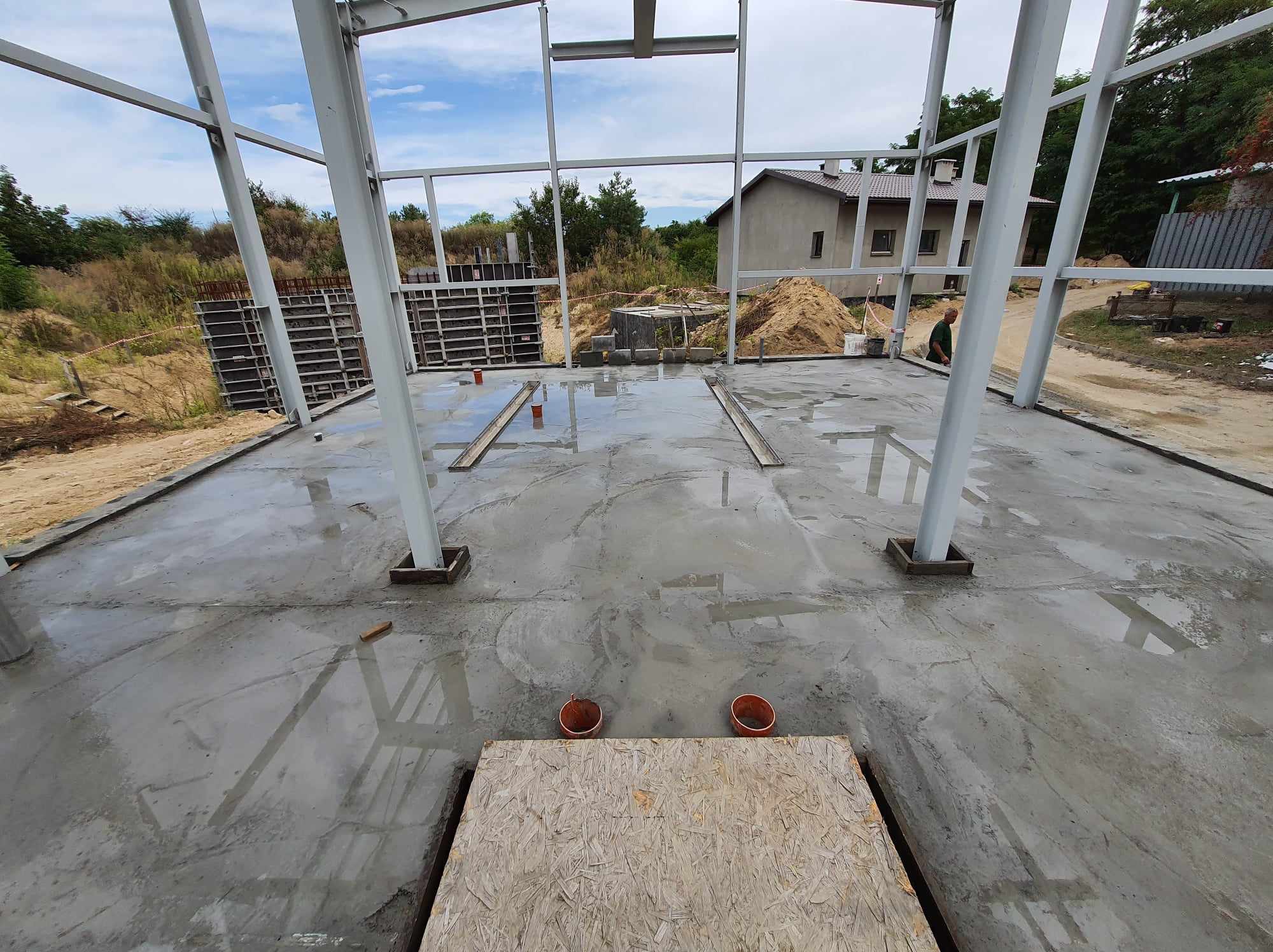 Wnętrze budowanej hali z widoczną konstrukcją stalową, mokrą betonową posadzką i deską na pierwszym planie, w tle widoczny budynek i teren.