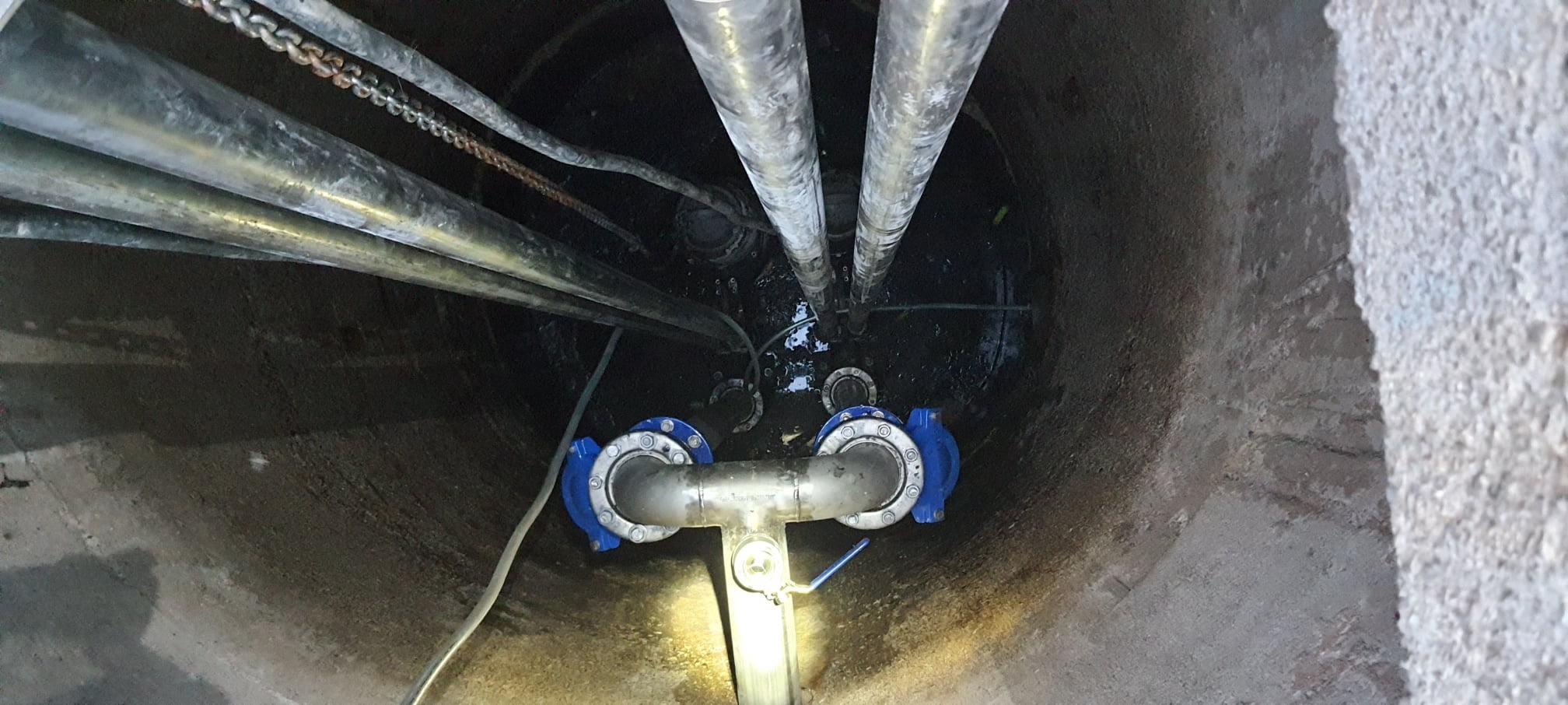 Wnętrze betonowej studni z rurami stalowymi, złączkami w kolorze niebieskim i przewodami, widok z góry.