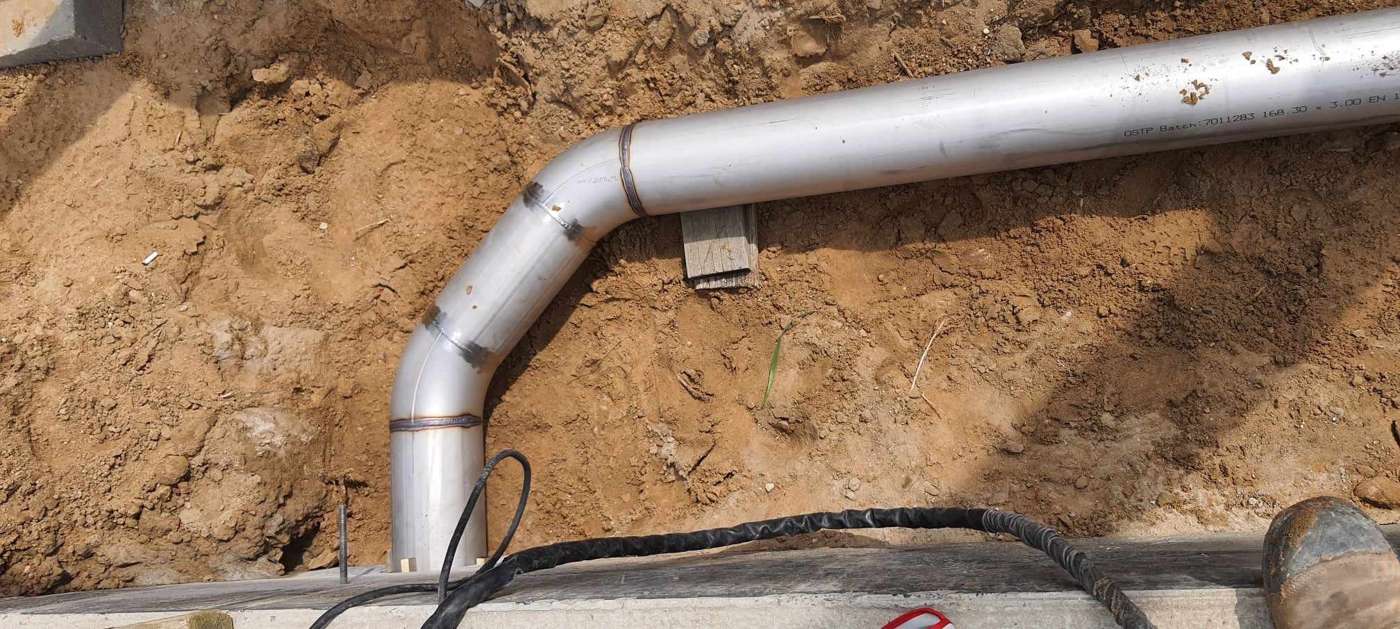 Spawany, nierdzewny element rurowy z kolankiem, wkopany w ziemię, Zawiercie. Widoczne spawy i oznaczenia producenta na rurze.