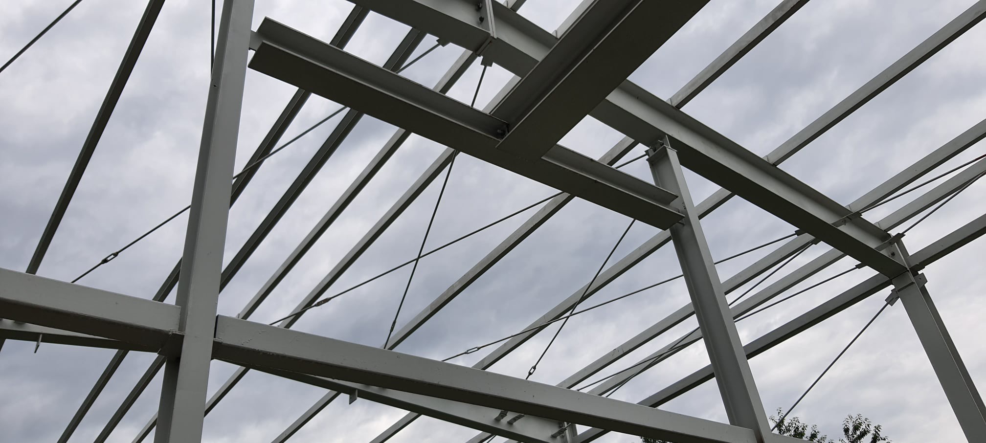 Szkielet stalowej konstrukcji hali, widok z dołu na krzyżujące się profile i liny mocujące na tle pochmurnego nieba.