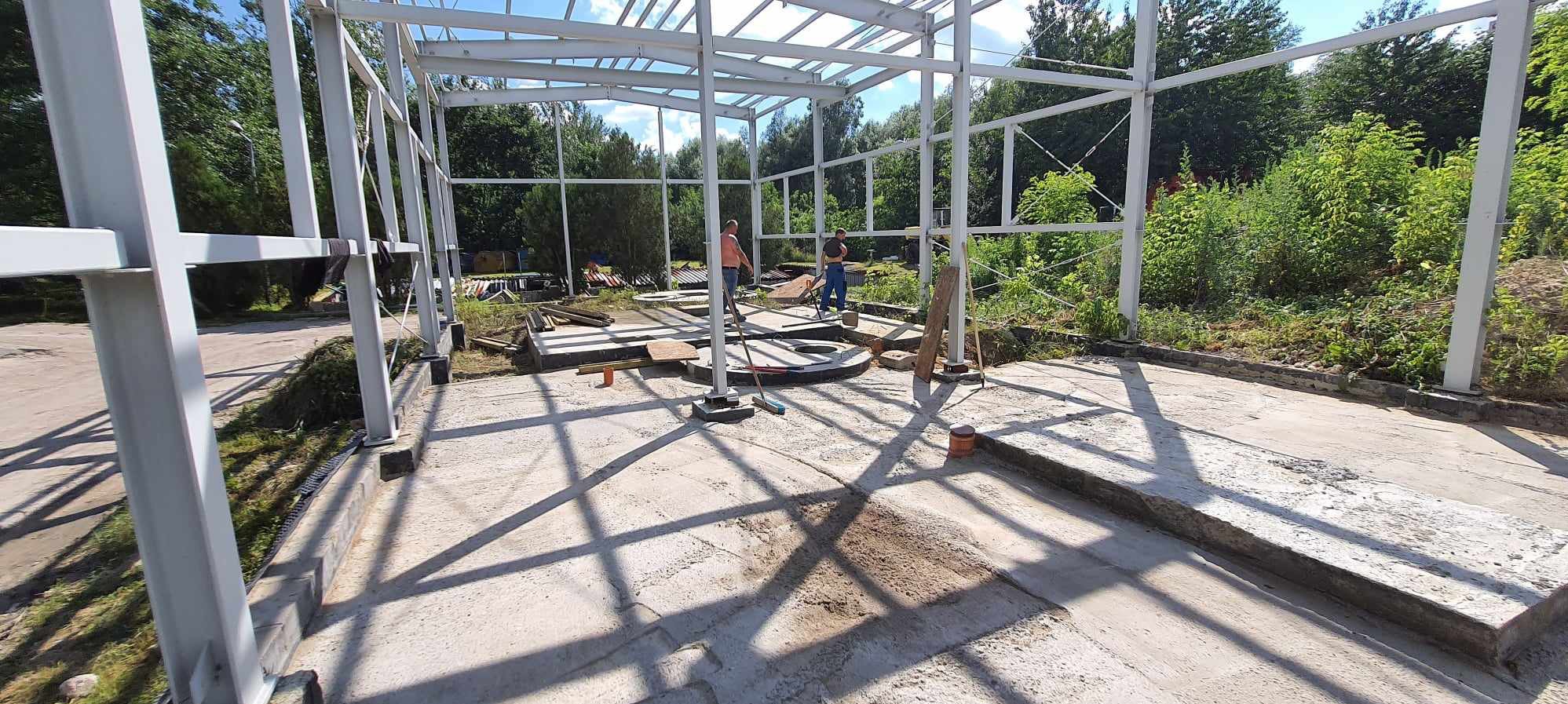 Wnętrze budowanej hali z widoczną stalową konstrukcją szkieletową, betonową posadzką w trakcie prac i dwoma pracownikami w tle.