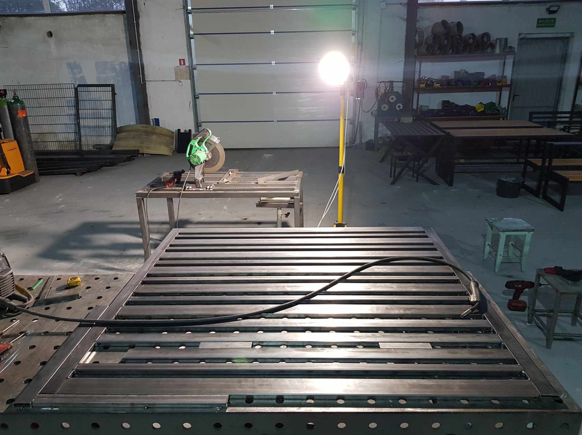 Konstrukcja metalowa z profili, prawdopodobnie rama, na stole spawalniczym w warsztacie, oświetlona lampą roboczą, z widocznymi narzędziami do obróbki metalu.