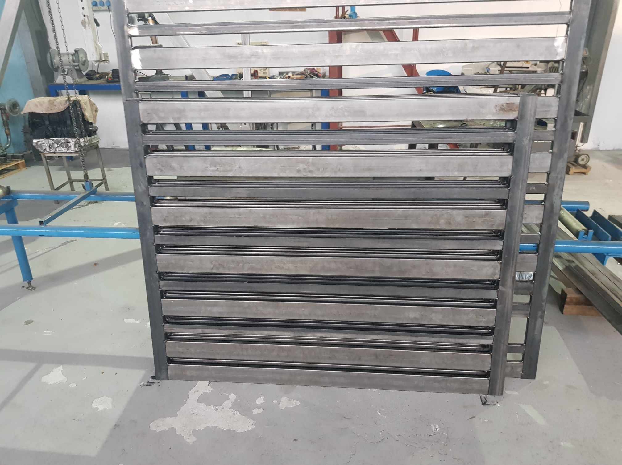 Surowy, metalowy panel ogrodzeniowy z poziomymi szczeblami i ramą, stojący w warsztacie, widoczne ślady spawania i obróbki.