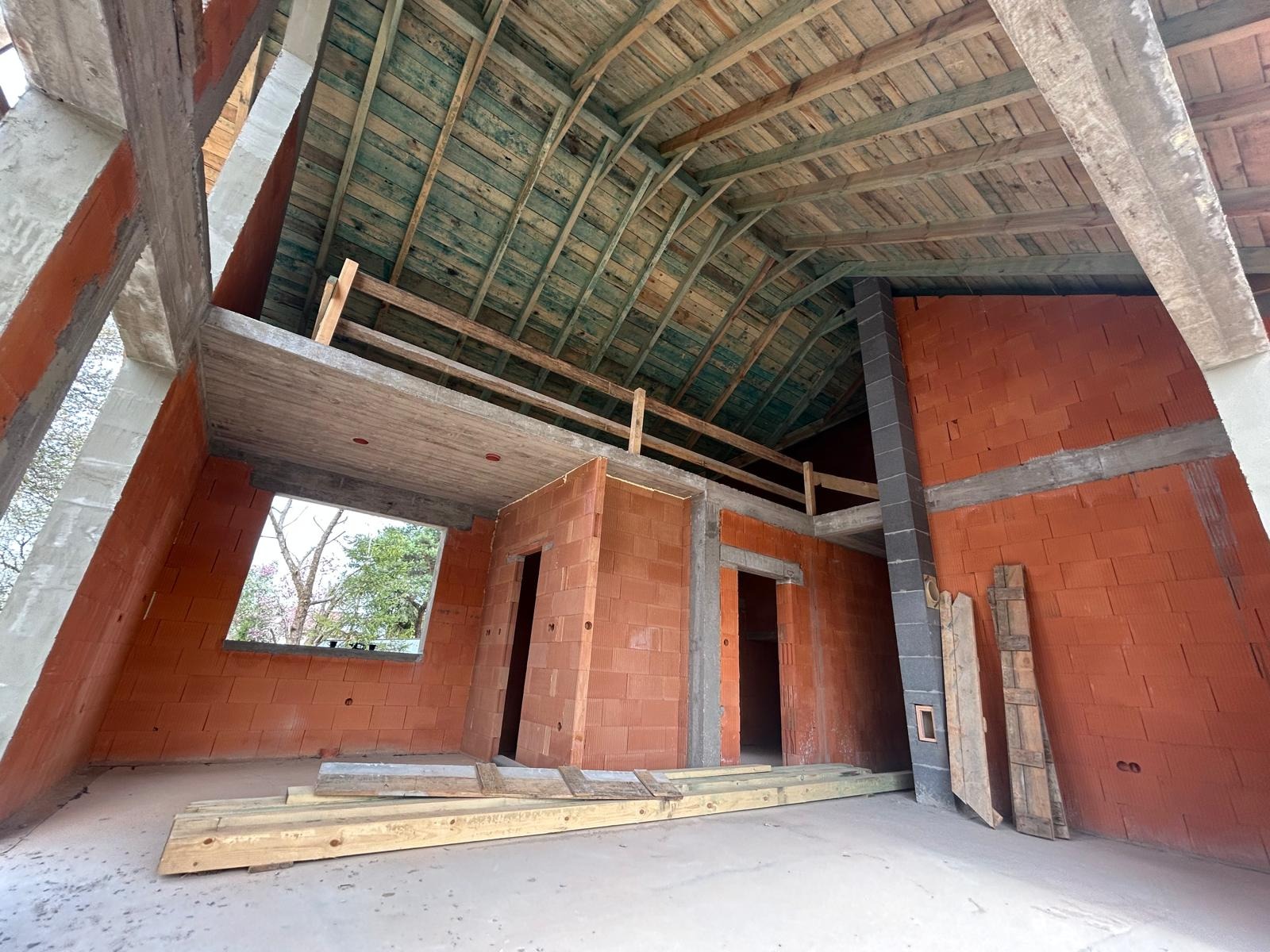 Surowy stan poddasza z ceglanymi ścianami, drewnianym stropem i betonowymi elementami konstrukcyjnymi. Widoczne otwory okienne i drzwiowe, fragment komina.