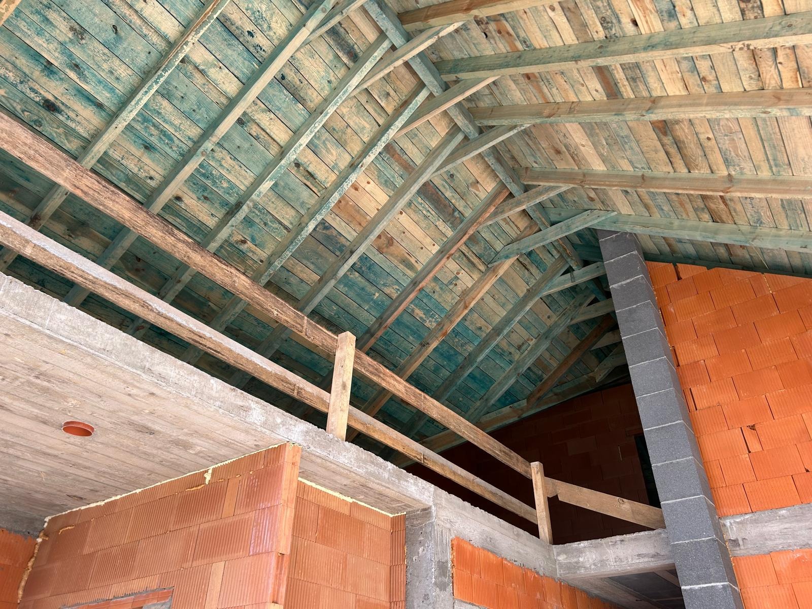 Surowy stan poddasza z drewnianą konstrukcją dachu, ceglanymi ścianami i betonowym stropem. Widoczne elementy instalacji kominowej i drewniane barierki.