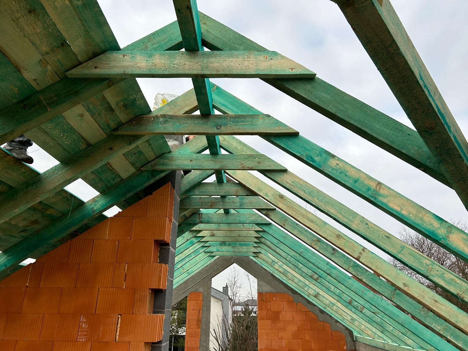 Konstrukcja dachu z zielonych belek drewnianych i ceglanymi ścianami w trakcie budowy. Widok od wewnątrz, na tle pochmurnego nieba i fragmentu budynku.