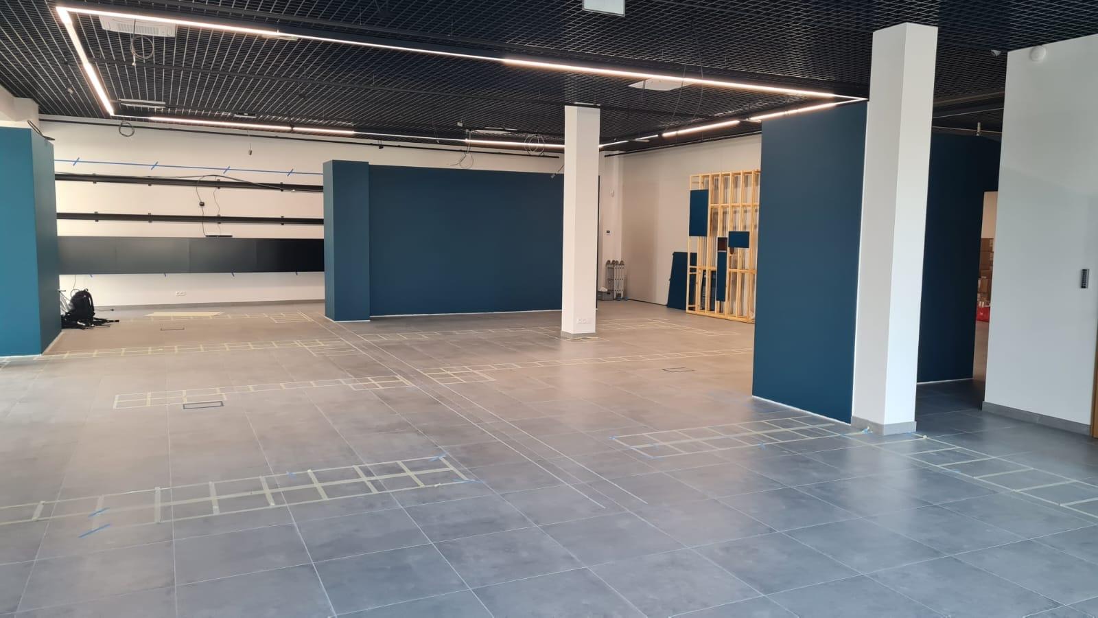 Wnętrze w trakcie wykańczania: ciemne ściany, szara podłoga z zaznaczonymi polami, białe kolumny. Widoczna instalacja pod sufitem i drewniana konstrukcja ścianki działowej.