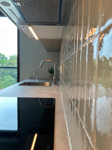 Nowoczesna kuchnia z błyszczącymi, pionowymi płytkami w odcieniach szarości, czarną płytą indukcyjną i stalowym zlewem pod oknem. Minimalistyczny design.