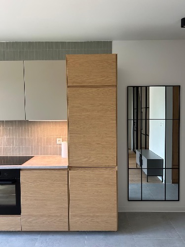 Nowoczesna kuchnia z drewnianymi frontami, szarymi płytkami i dużym lustrem w czarnej ramie, odbijającym wnętrze. Minimalistyczny design, jasne kolory.