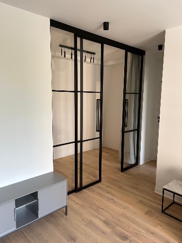 Nowoczesne, przesuwne drzwi szklane z czarnymi szprosami, oddzielające przestrzeń. Jasne panele podłogowe i minimalistyczne meble w tle. Oświetlenie sufitowe.