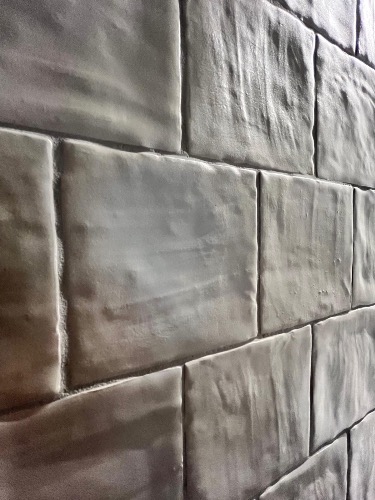 Ściana wyłożona szarymi, prostokątnymi płytkami ceramicznymi z wyraźną fakturą, ułożonymi w regularny wzór. Widoczne fugi. Ujęcie pod kątem.