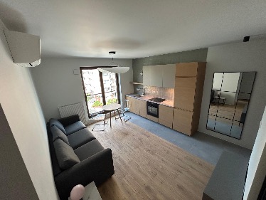 Jasne, nowocześnie urządzone studio z drewnianą podłogą, szarą sofą i zabudową kuchenną z drewnianymi frontami. Widok na balkon i miasto w tle.