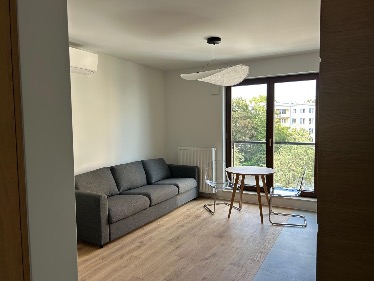 Jasne wnętrze salonu z szarą sofą, okrągłym stolikiem i widokiem na zieleń za oknem. Minimalistyczna lampa sufitowa i drewniana podłoga dodają przytulności.