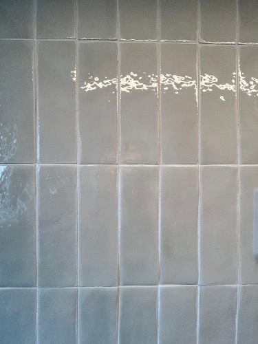 Błyszczące, pionowo ułożone, szare płytki ścienne w łazience, z widocznymi refleksami światła. Minimalistyczny design, fuga w odcieniu zbliżonym do płytek.