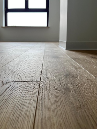 Ułożona podłoga z paneli w odcieniach brązu, widok z niskiej perspektywy, z oknem w tle. Minimalistyczne wnętrze z listwami przypodłogowymi.