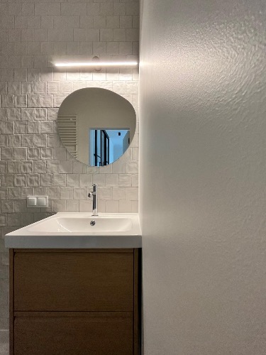 Nowoczesna łazienka: biała umywalka z drewnianą szafką, lustro o nieregularnym kształcie, oświetlenie LED i ściana z białej, strukturalnej cegły.