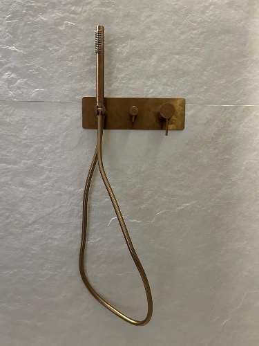 Mosiężna bateria prysznicowa z deszczownicą na tle jasnoszarej, strukturalnej ściany. Minimalistyczny design, widoczne detale wykończenia.