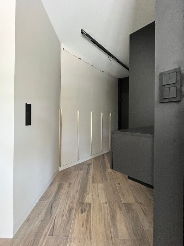 Nowoczesny przedpokój z drewnopodobną podłogą, zabudowaną szafą z złotymi uchwytami i minimalistycznym oświetleniem szynowym. Ciemne akcenty w postaci drzwi i włączników.